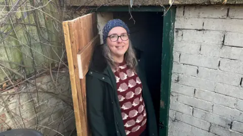 Mary Andrews has light brown, shoulder length hair and is wearing a blue bobble hat which covers her head. She has brown-frame glasses and a dark green overcoat over a brown woollen jumper with white hedgehog designs on it. She is standing in the open doorway of a small air raid shelter with a new wooded door with a green doorframe, some bare shrub branches are growing on a wooden fence.