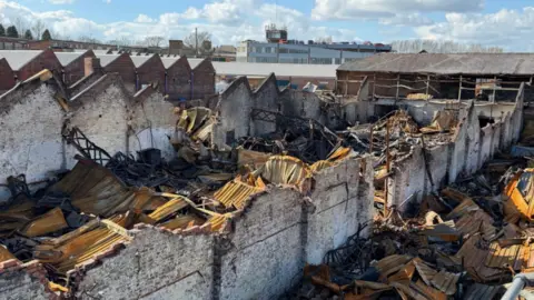 A damaged factory consisting of rows of pitched white-washed walls, with its roof caved in. Extensive debris can be seen on the floor of the building. It is a sunny day.