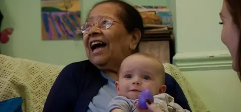 An elderly woman holding a baby. SHe has glasses and dark hair and is looking to the side with her mouth open smiling. The baby is looking at the camera.