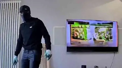 A man dressed in black with a balaclava standing in a living room with the TV on behind him.