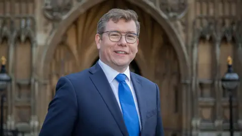 PA Media Peter Gibson, chief executive of Fighting with Pride, standing outside the Houses of Parliament, wearing a navy suit and a royal blue tie. He has brown hair and glasses.