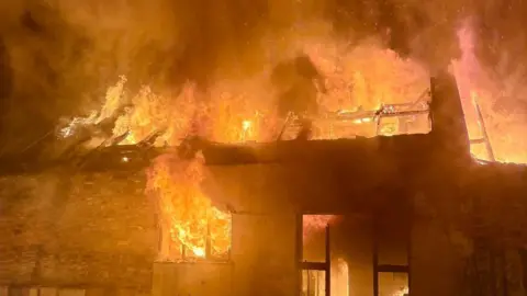 A house on fire. Flames are issuing out from the doors, windows, and the roof.