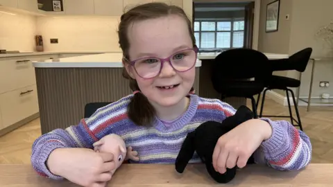 BBC, Leanne Rinne A young girl smiling in a striped purple jumper wearing glasses and holding to soft toys in her hands. She is sat in a kitchen. 