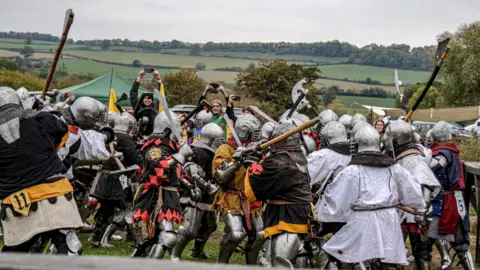 Large outdoor medieval battle with dozens of participants wearing full metal armour and helmets packed closely together. Many hold swords, axes, or polearms raised mid-fight, creating a dense cluster of movement. Spectators stand behind the fighters, some holding up phones to record. The setting is a grassy field with rolling green countryside and trees in the distance under an overcast sky. 