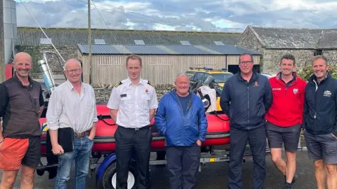 A group of male Fleet Bay Inshore Rescue Service trustees are standing in front of their current rescue boat alongside a representative from HM Coastguard