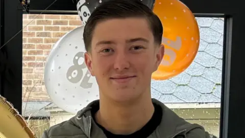 An 18-year-old man standing in front of orange, white and black balloons by a window. He has short dark hair and is smiling. Wearing a blue jacket and a black top underneath.