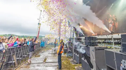Kubix Kubix festival's stage photographed from the side. A security guard is standing in front of it as pink and yellow confetti flies in the air with stage smoke going off. Fans are lined behind a metal barrier and have their hands up in the air.