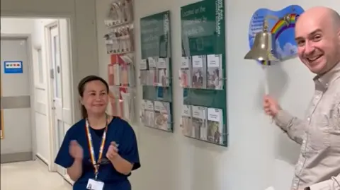 Ben Goldsborough Ben Goldsborough is on the right hand side of the picture ringing a wall-mounted bell in a hospital. To the left a female NHS worker is clapping.