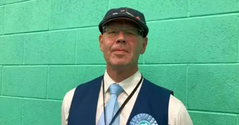 LDRS A man in a white shirt with blue waistcoat and blue cap and light blue tie and light blue Reform UK rosette standing against a turquoise wall