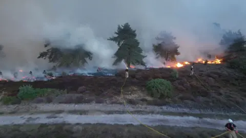 Ryan Lovejoy. Los bomberos, con mangueras, combaten un incendio forestal en Dunwich Heath. Entre la densa humareda y las cenizas, se ven algunos árboles verdes dispersos.