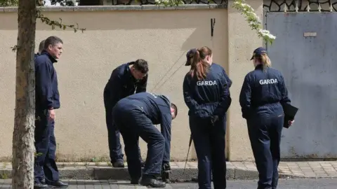 PA Media Seven uniformed Garda officers in navy conduct a search of a pavement outside a house. There is a cream wall behind them and a grey metal fency. There is the trunk of a tree in the left of the picture.