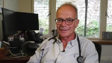 Dr Opher smiles into the camera. He is sitting in a GP consulting room with a computer and desk behind him. He is sitting in a desk chair. He has a stethoscope around his neck and is wearing glasses and a white shirt.