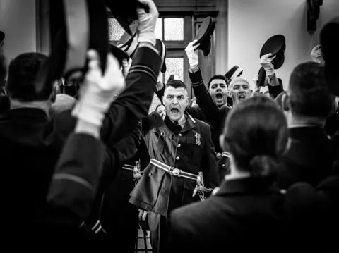 Andrew Wheeler A black and white image of officers holding their caps in the air as a man centre stage walks down the isle.  The three officers facing the camera are either cheering or shouting and the photo captures the moment.