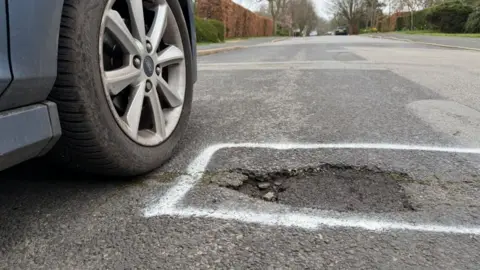BBC A pothole in a quiet road marked in white paint next to a car wheel. The pothole is an uneven shape and is sunken into a road which is elsewhere patched up in different areas.