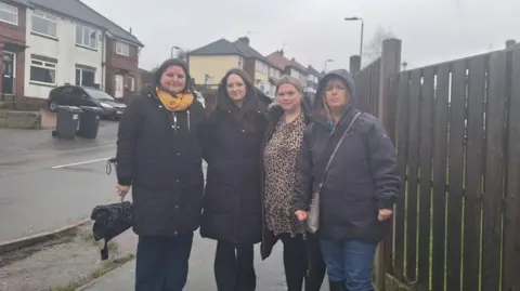 Four women stood close together in a row. They are wearing jackets, with one woman who has her hood up. They are stood on the pavement of a road with a fence and houses in the background. 