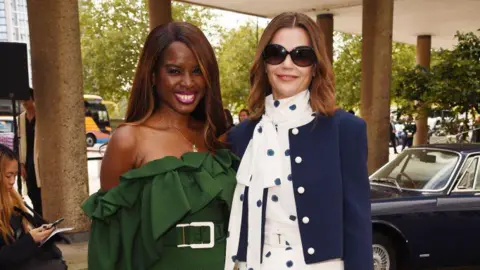 Getty Images June Sarpong wearing a green off-the-shoulder dress with a large belt. She is standing next to Victoria Starmer who is wearing a navy jacket with white buttons and a white dress with blue spots. 