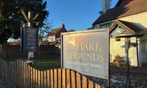 A pub site with a wooden fence in the foreground. There are two boards near the fence and a building is behind them.