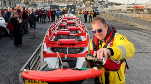 A man from the RNLI wearing a bright yellow jacket and a life vest pours a can of Guinness onto the tip of a gig point. 