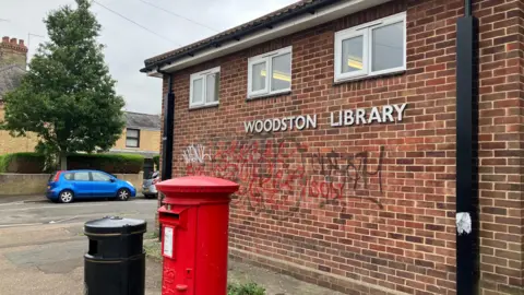 Outside of Woodston library, there is a sign in capital letters and some graffiti on the building.