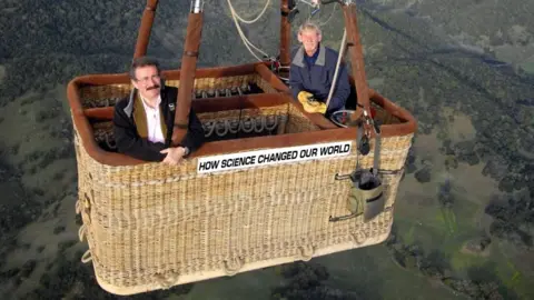 Lord Winston with dark hair and beard, wearing a dark jacket and white shirt, resting his arms on the basket of a hot air balloon in flight. Another man wearing a blue jacket is at the other side of the basket (presumably the pilot). He is wearing yellow gloves. The balloon is travelling over fields and trees.