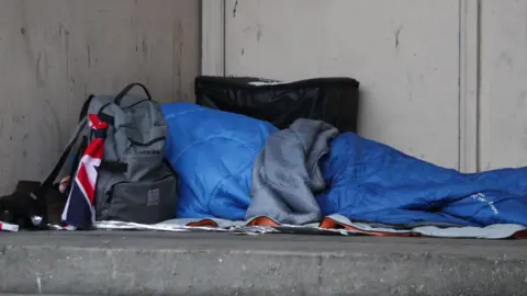 Rough sleeper in blue sleeping bag next to a rucksack on hard concrete floor
