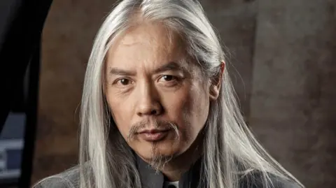 Clem So with white long hair and brown eyes with a small goatee beard. He is wearing a grey suit. 