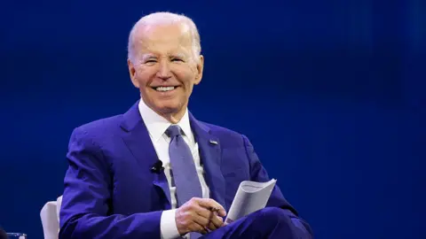 Getty Images Joe Biden speaks at the Society for Human Resource Management (SHRM) Annual Conference at the San Diego Convention Center on 2 July 2025 in San Diego. He is wearing a blue suit and tie and white shirt. He has grey hair and is smiling, holding a pen and paper.