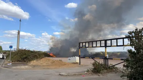 Will Jefford/BBC A field where a hedge is on fire and thick smoke billows into the air. The photo is taken from a road at the side of the field. There is a black and yellow barrier preventing cars from driving into the field.