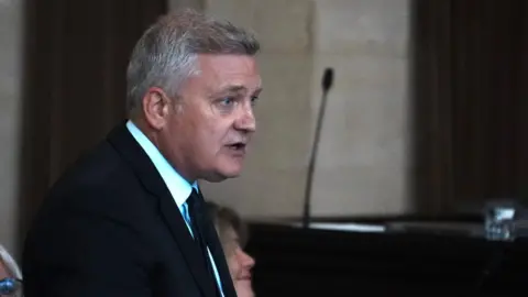 John Fairhall/BBC Deputy council leader Russell Quirk, who is wearing a suit and has short grey hair, speaking during a meeting.