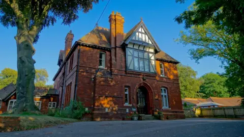 Bernadette House Brick building with Tudor frontage around first floor window. There are a further four windows in the front and a door. There are trees surrounding the property and a tarmac drive in front.