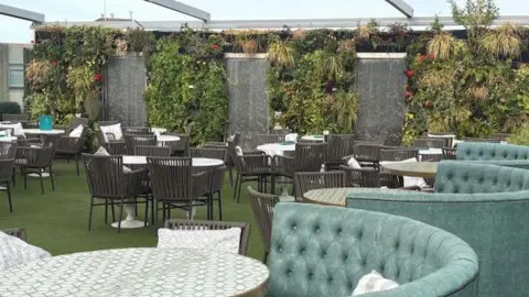 Roof area of a building being used as a restaurant. In the foreground is a turquoise-coloured high-backed chair in a crescent shape, with a table. Behind are a number of tables and chairs. There is a green carpet. The sky is visible.