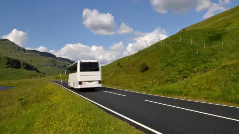 A white coach drives along a road with grassy verges on each side and a mountain in the background. 