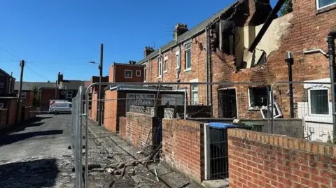 Fencing surrounds the destroyed house with explosion debris and one of the house windows hanging 