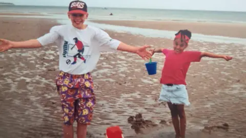 Alicia Manners A young boy and girl smiling stood with their arms stretched open on a beach. The girl is holding a plastic bucket and is wearing a red t- shirt and a denim skirt. The boy is stood next to a plastic bucket and is wearing colourful patterened shorts and a t-shirt with 'Puma' written on. 