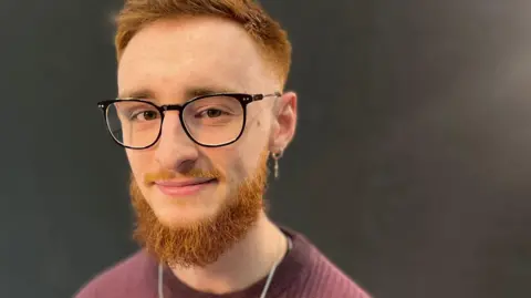 Ginger haired man with a beard and a half-smile, wearing a burgundy ribbed jumper and metal neck chain and large round glasses sitting in front of a black background.