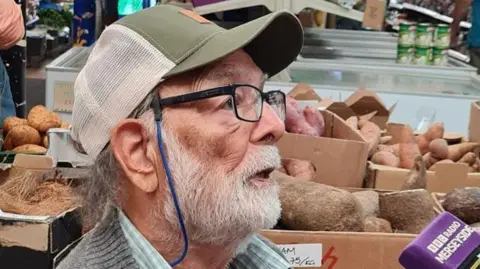 Kirpal Matta, pictured in 2024, has a grey beard and is wearing black rimmed glasses and a green-and-white baseball cap. He is also wearing a green checked shirt and a grey cardigan.
