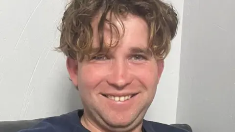 James Nunan has long, curly brown hair drooping downwards, partially covering one eye. He is smiling in front of a white wall and wearing a navy T-shirt.