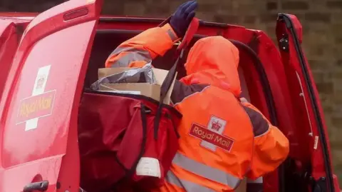 A Royal mail worker in an orange coat with a hood up loads a bag of parcels out of a van. 