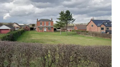 A rural or semi-rural residential scene with a large open grassy field in the foreground. The field is unevenly cut and bordered at the front by a low hedge made of thin, leafless branches, forming a natural boundary along the roadside or edge of the property.
In the middle of the image, there is a two-storey red brick house positioned centrally within the field. The house has a symmetrical front with white-framed windows and a central front door. The roof is dark grey and slopes gently, with chimneys visible on either side. Behind and beside the main house, there are additional outbuildings and structures, including what appears to be a barn or garage with a wide opening.