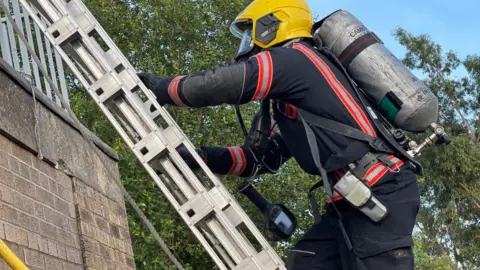 Cambridgeshire Fire and Rescue Service A firefighter wearing a black fire suit, with a yellow helmet and an oxygen cylinder on his back, climbing up a silver ladder