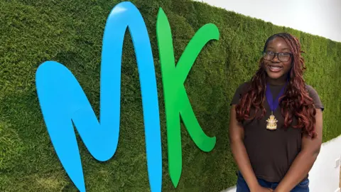 A young woman wearing glasses and a ceremonial badge stands with folded hands beside a large blue-and-green logo attached to a textured green wall, with office space visible in the background.