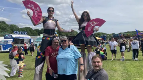 STUART PRANDLE/BBC A photo from the festival-style Pride celebrations at The Sands in Durham. A field with food trucks and fairground rides stretches out behind a group of people posing - two are wearing tall stilts and waving pink fans with the word "whatever" across them.