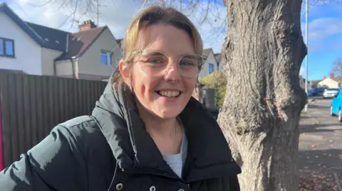 Kate Bradbrook/BBC Sam smiles at the camera, standing on the pavement next to a tree with houses behind her. She wears clear glasses and a black puffer coat over a grey T-shirt.