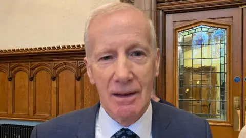 Gregory Campbell blue jacket, light-coloured shirt and dark floral tie in the main hall of the Guildhall. Wood panelling can be seen behind him as well as a wooden door with stained glass window.