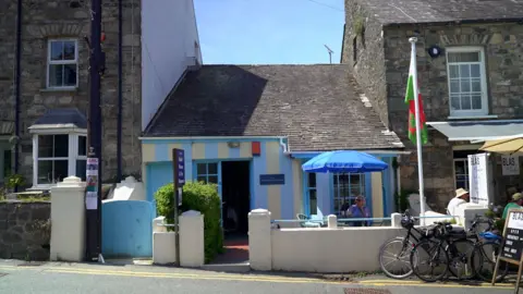 The view from the other side of the street of the single story dolls house museum. It sits with a grey slate roof sandwiched between two grey stone brick houses. The outside of the museum has been painted with a thick white and blue stripe pattern and a table with a blue parasol is positioned outside beside a welsh flag pole.