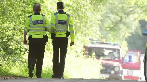 Sussex News and Pictures Two police officers stood in a country road with emergency service vehicles.