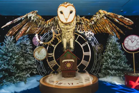 Rise Productions A scene from a magical grotto, with several clocks of different shapes and sizes, including a cuckoo clock, and a large model of an owl swooping over the landscape of snowy trees