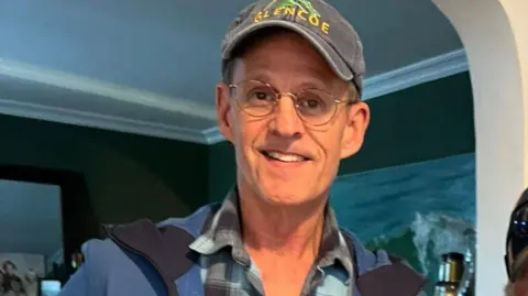 Facebook Brian McGillicuddy has round glasses and is wearing a Glencoe baseball cap, pale blue and black checked shirt and blue soft shell jacket.