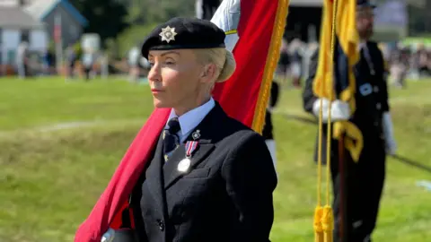 McCabe in uniform carrying the Manx flag. There is green grass around and other flag bearer standing to attention.