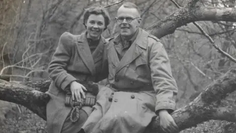 Éanna Mac Cana/Little Ease Films A black and white image of Dr Denis Burkitt and his wife Olive sitting on a large tree branch, smiling at the camera. Sitting on the left, Olive has on a large winter coat and is holding a striped handbag. Her hair is pinned back into an up do. Denis is wearing a long trench coat and has short hair. He is also wearing a pair of circular glasses.
